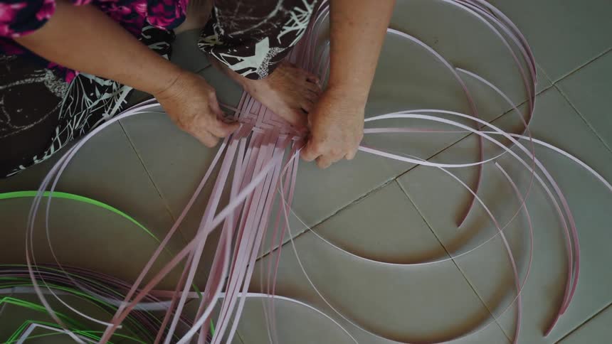 A person is weaving or braiding multiple strands of pink and green plastic strips on a tiled floor