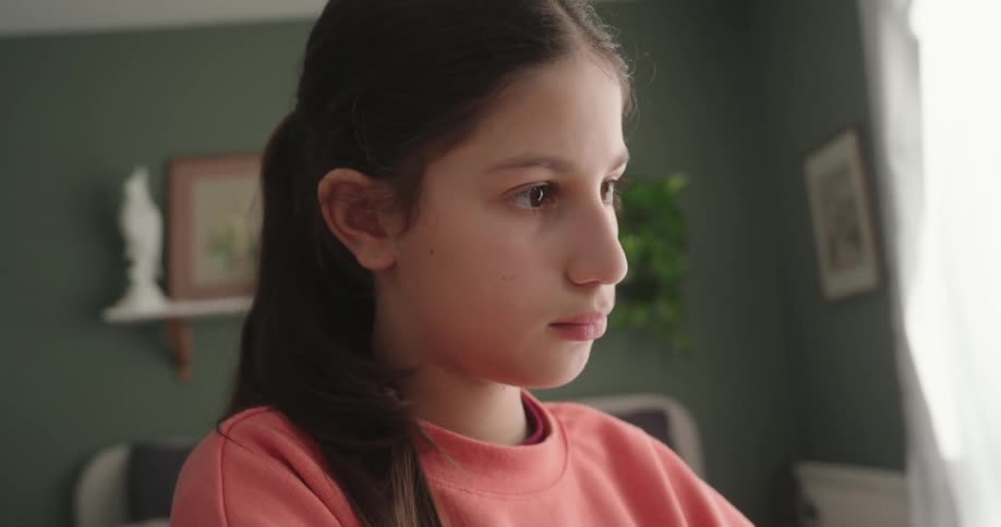 A girl wearing a pink sweatshirt is typing on a laptop in a room with a green wall and a window with white curtains