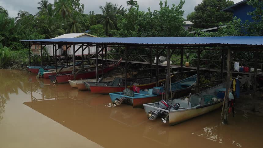 Several small boats are lined up under a blueroofed shelter beside a muddy brown river surrounded by lush green foliage