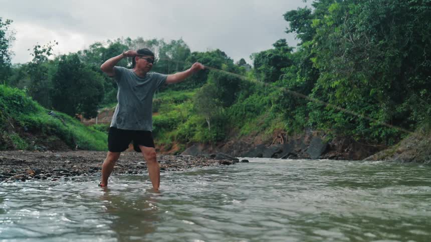 Standing in shallow water a man powerfully throws a fishing net in a wide arc initiating the traditional fishing technique of casting a net to capture fish in a river surrounded by lush green trees under an overcast sky