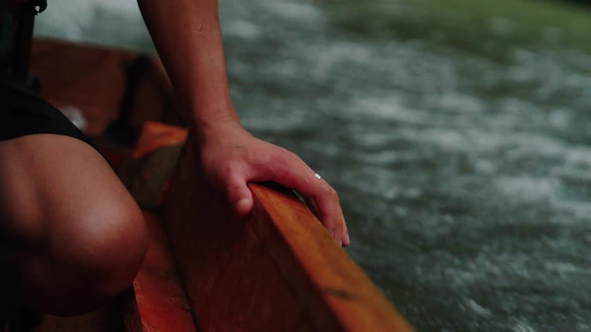 A person is canoeing down a river holding the canoe edge with their hand as the water flows quickly by