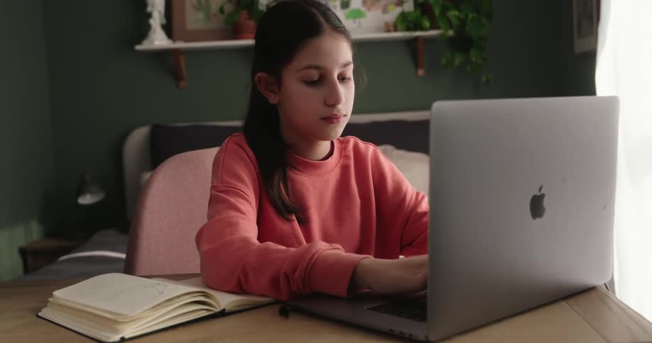 A girl wearing a pink sweatshirt is typing on a laptop in a room with a green wall and a window with white curtains