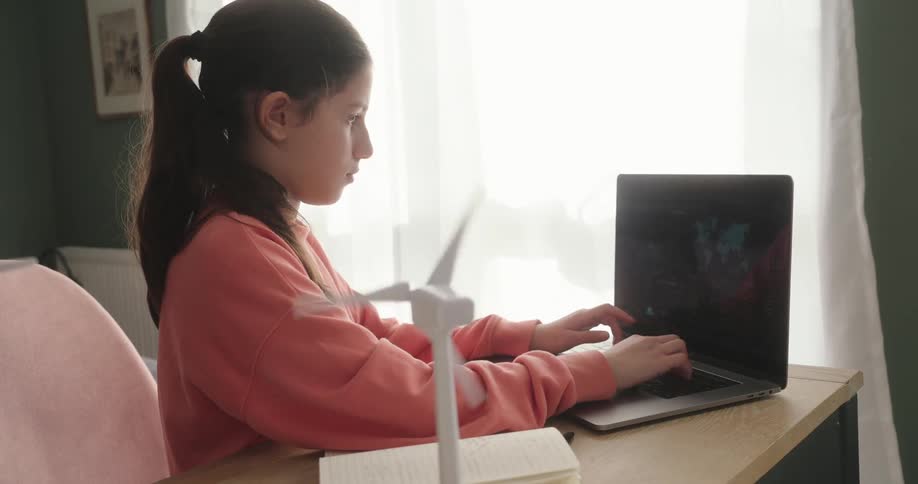 Girl in a pink blouse works concentratedly on a laptop sitting at the table with a model of a wind turbine next to it