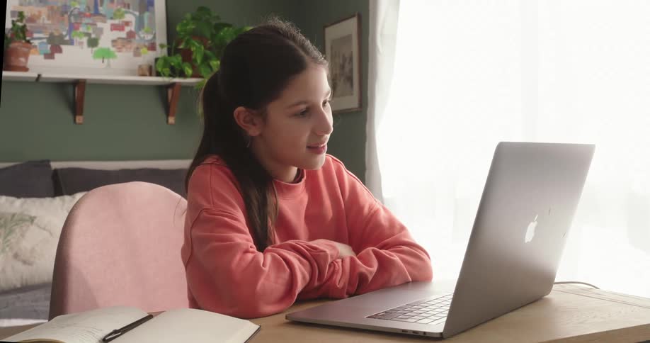 A teenage girl sits at herdesk in front of a laptop and waves