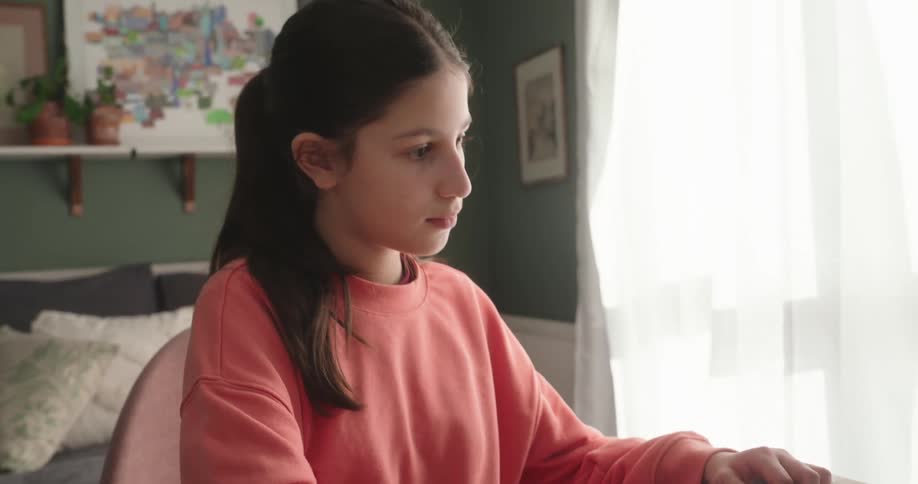A girl wearing a pink sweatshirt is typing on a laptop in a room with a green wall and a window with white curtains
