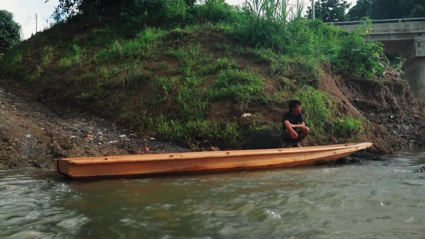 With a gentle river flowing in the foreground and a bridge visible in the right background a man is captured squatting on a simple wooden boat beached on the riverbank against a backdrop of lush greenery waiting for riders