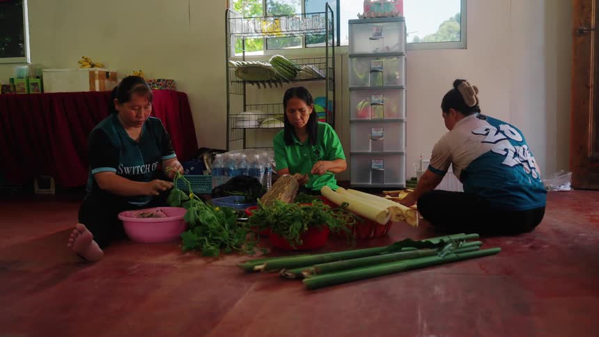Three women sitting on the floor of a living room working with bamboo and vegetables in colorful bowls