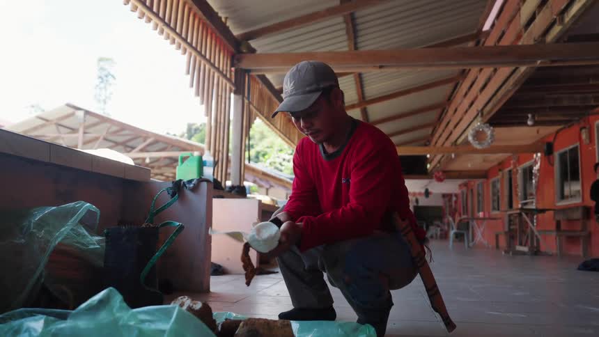 A person wearing a red shirt and a cap is crouching on a tiled floor in an open market peeling tubers over green plastic bags