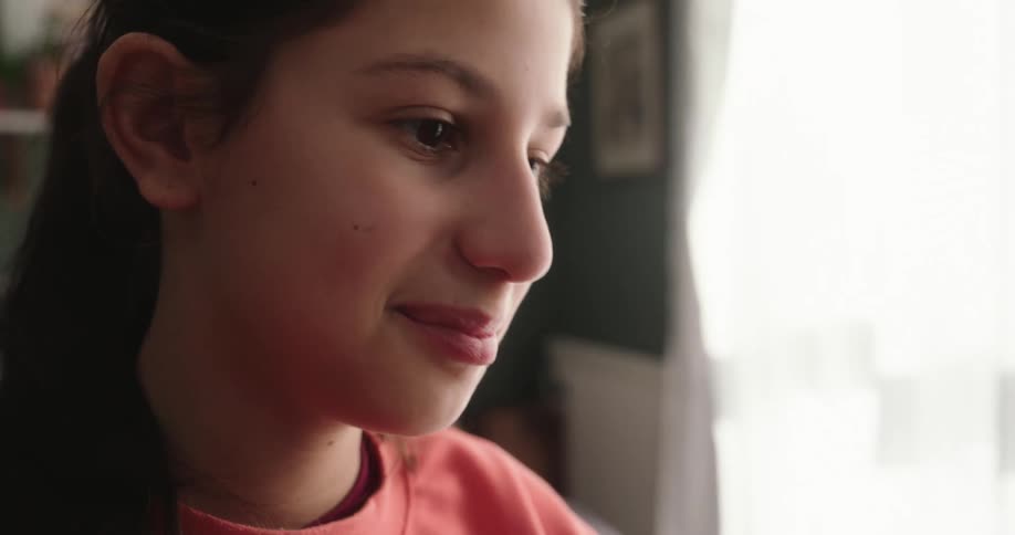 A girl wearing a pink sweatshirt is typing on a laptop in a room with a green wall and a window with white curtains