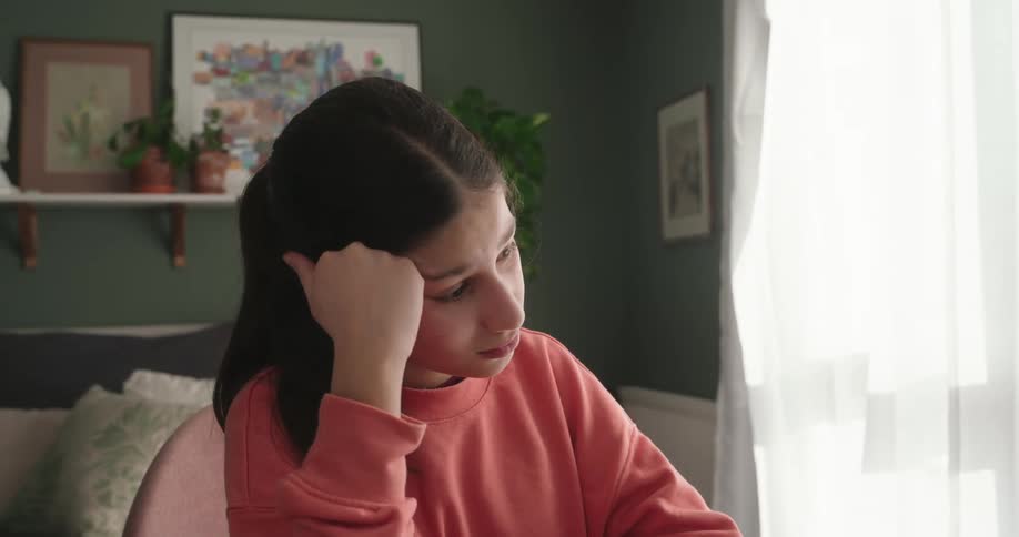 A girl wearing a pink sweatshirt is bored in front of a laptop in a room with a green wall and a window with white curtains
