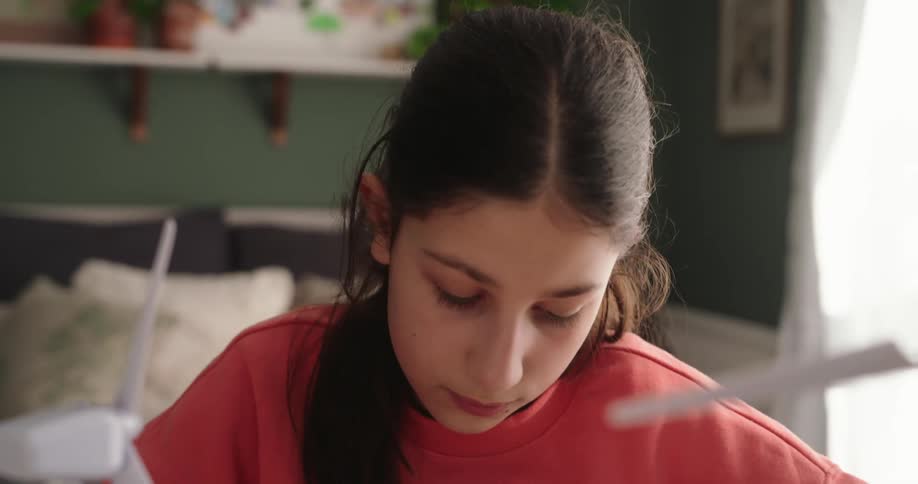 A girl looks around and puts a pen to her face as she thinks and looks at a book she has open on the desk