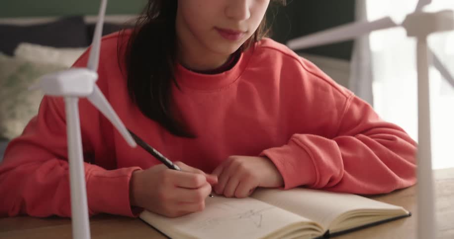 A girl wearing a pink sweatshirt is drawing wind turbines on a notebook inspired by two small scale models on her desk in a room with a green wall and a window with white curtains