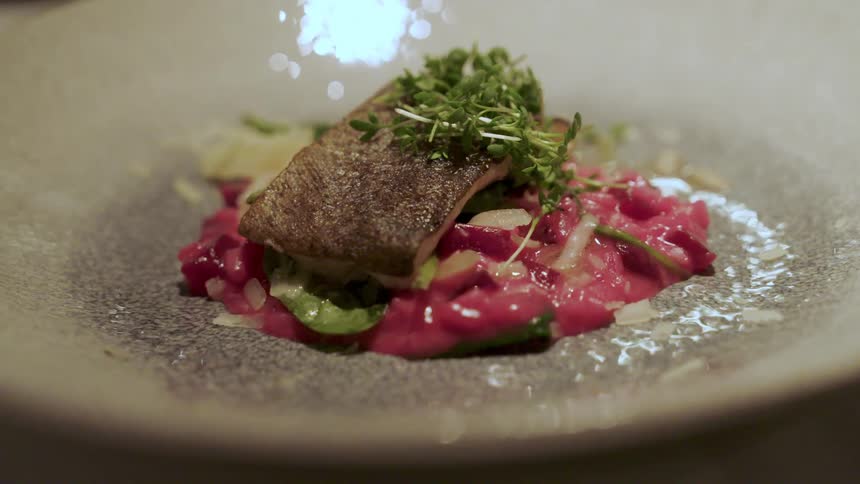 A closeup shot of a plate presents a piece of cooked fish with crispy skin atop a bed of beetcolored risotto and leafy greens