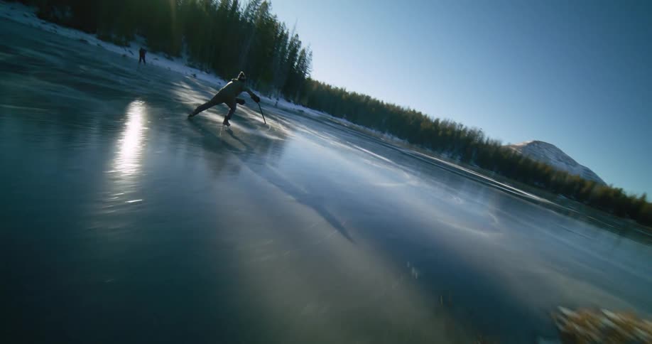 Hockey players skate across a frozen pond surrounded by a conifer forest on a sunny winter day