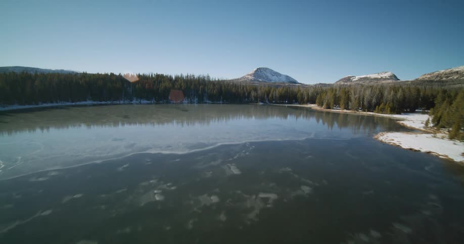 Partially frozen lake reflects the blue sky surrounded by a dense coniferous forest and snowcapped mountains in the background