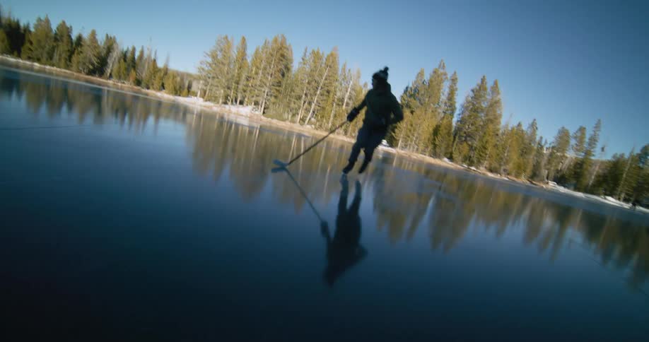 Hockey player ice skates backward on a frozen pond surrounded by conifer forest on a winter day