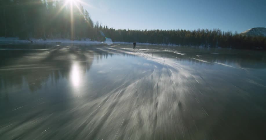 Two people are ice skating and playing hockey on a partially frozen lake surrounded by snow and pine trees on a sunny day