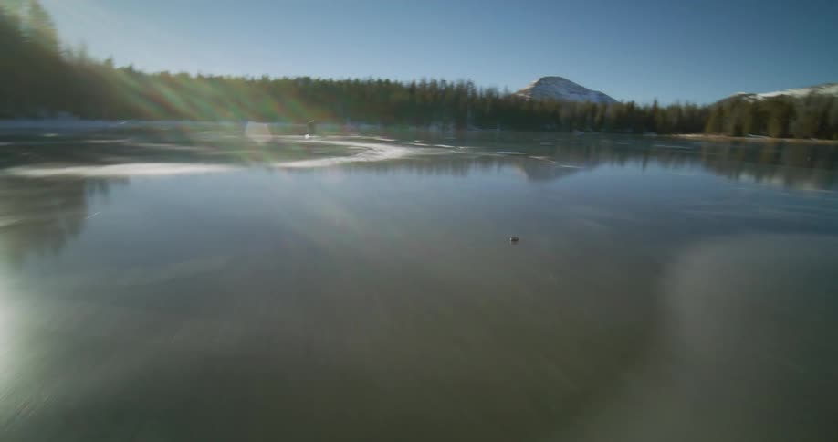 Two hockey players slide on a frozen lake reflecting the sunlight with mountains and trees in the background