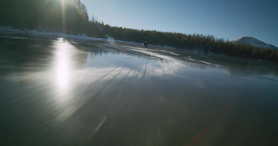 Person skates and plays hockey on a frozen lake reflecting the sun surrounded by forest and mountains in the background