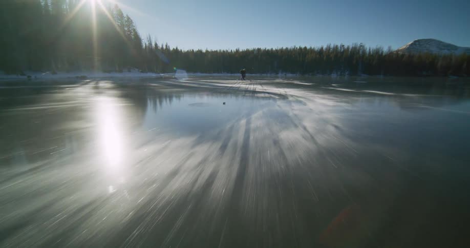 Hockey players play together on the reflective ice of a pond surrounded by conifer forest on a sunny winter day