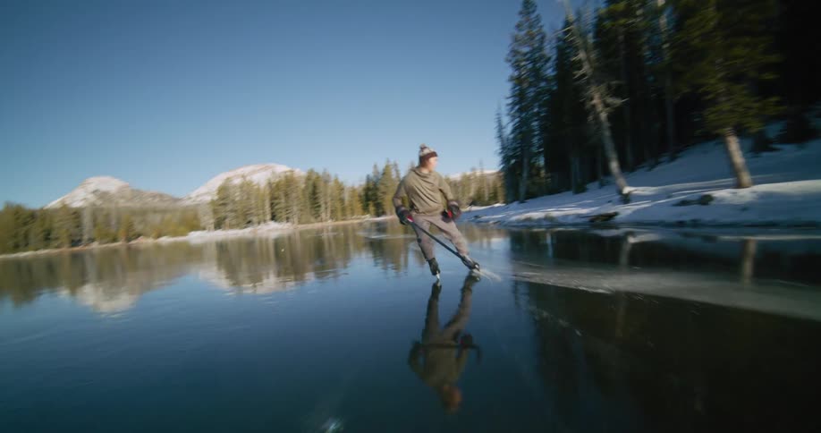 A person wearing a hat and coat skates across a frozen lake with a hockey stick and snowy mountains and tall thin trees visible in the background under a clear blue sky