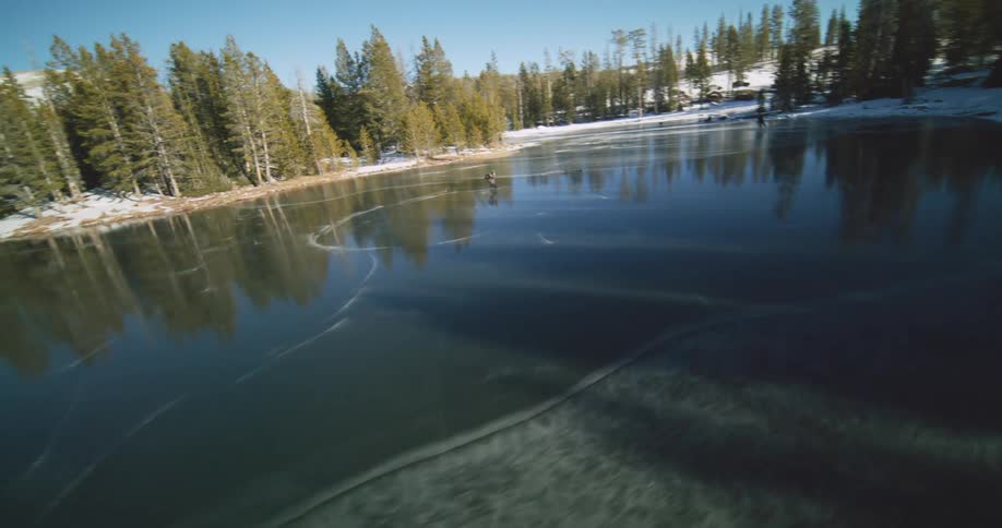 An ice skater is playing hockey on a frozen lake surrounded by a snow covered shoreline and towering trees under a bright sun