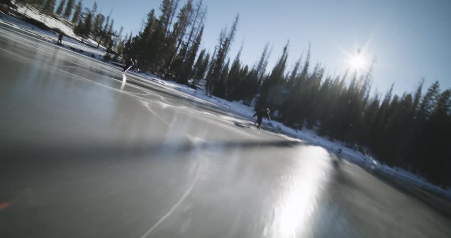 Person skating on a frozen lake amidst a natural landscape surrounded by trees and snowy mountains under a clear blue sky
