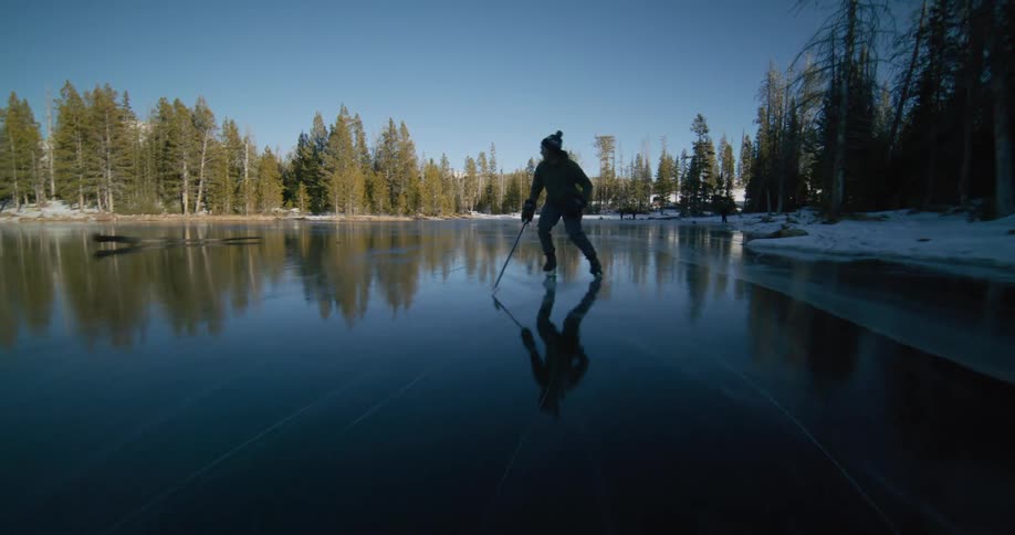 A person is skating backwards with a hockey stick on a frozen lake surrounded by a forest of evergreen trees under a clear blue sky