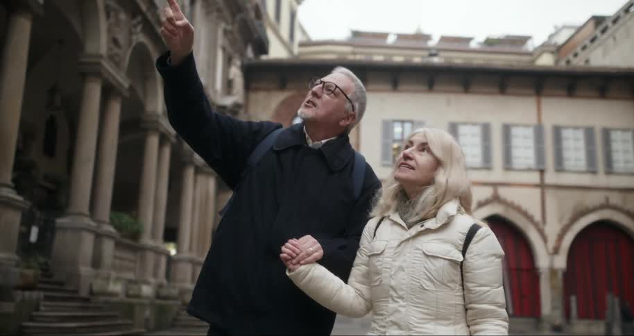 An elderly couple holds hands and looks upwards in a historic european courtyard