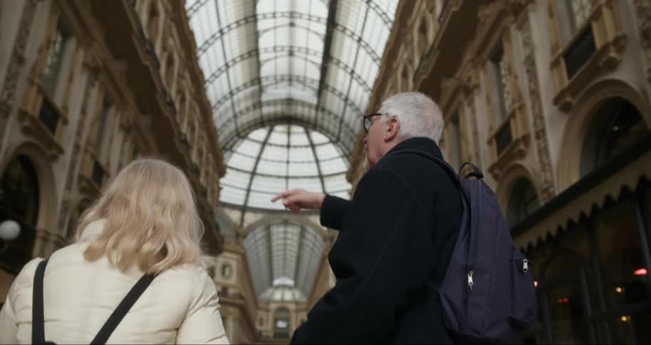 An older man with glasses wearing a dark coat and a backpack pointing towards the architecture inside an elegant glass ceiling gallery while a woman with blonde hair in a lightcolored coat listens attentively