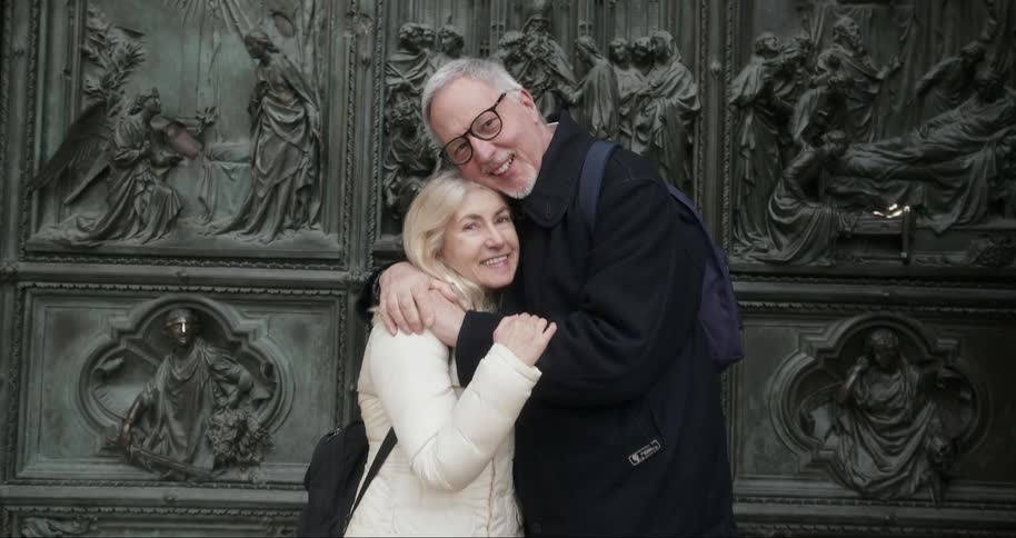 An elderly couple embraces warmly in front of an ornate historic bronze door adorned with intricate religious sculptures exuding love and companionship
