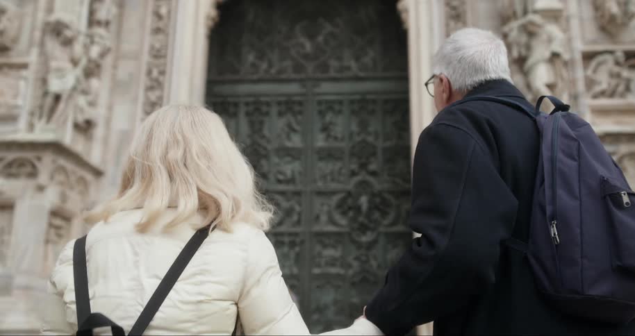 Older couple holds hands and smiles while walking towards an ornate cathedral decorated with stone carvings