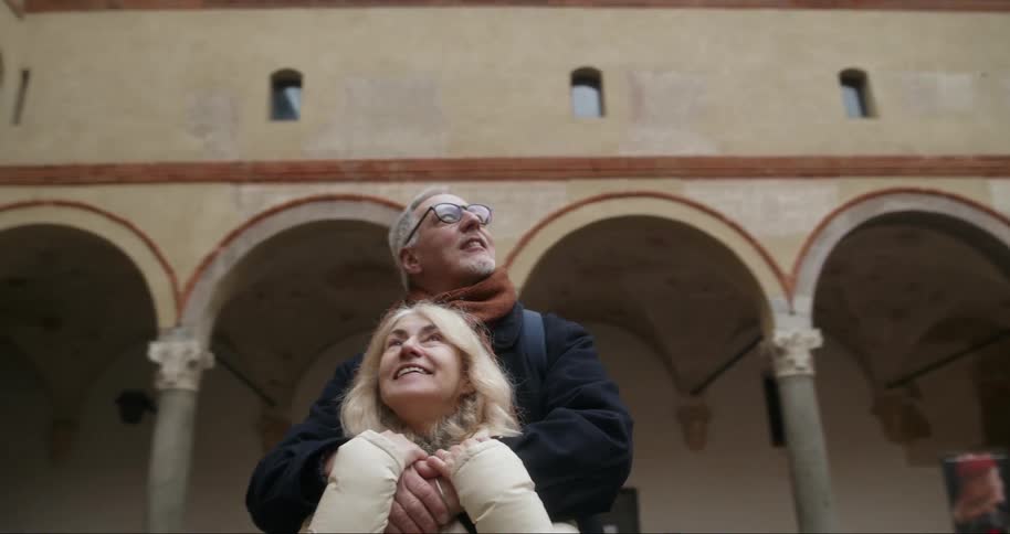 A man and a woman are hugging and looking happily around where there is a large and old building