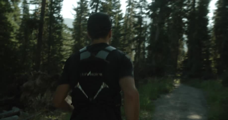 Man from behind running along a trail surrounded by tall trees with sunlight filtering through the leaves