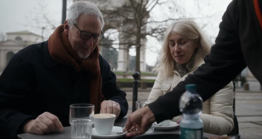 An elderly couple sitting at an outdoor cafe table on a chilly day dressed warmly smiling as they receive drinks from a server with a cozy and relaxed atmosphere