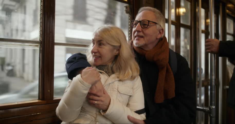 An elderly couple sits closely together on a vintage tram with the man lovingly embracing the woman from behind as they gaze out the window with warm expressions