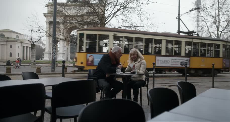 An elderly couple enjoys a tender moment at an outdoor caf sharing food and drink as a vintage yellow tram rolls by