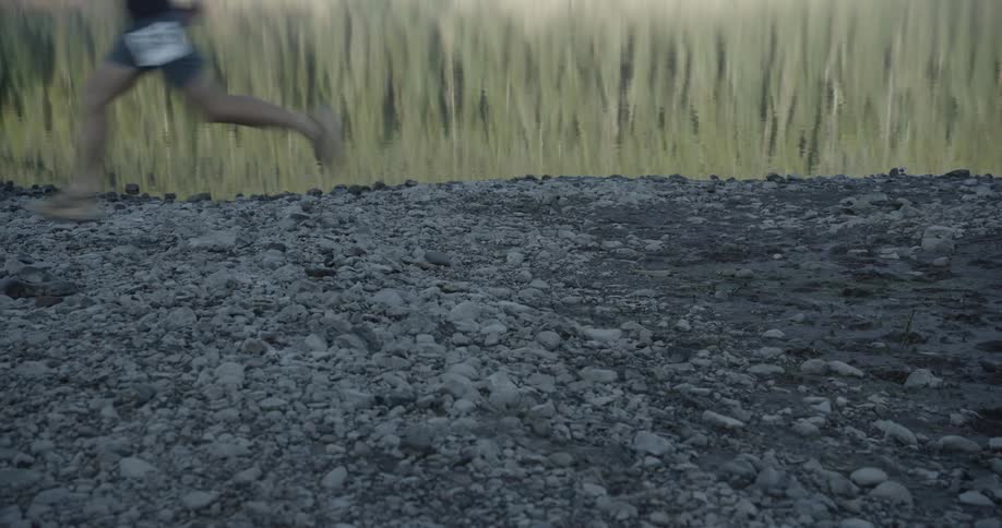Person runs barefoot on a rocky terrain at the edge of a lake with trees reflecting in the calm water