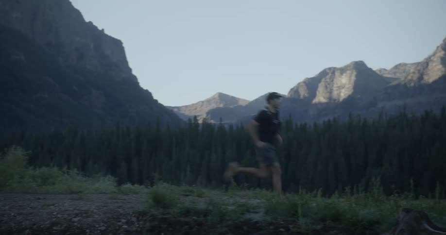 A man runs on a trail with a forest and towering mountains in the background