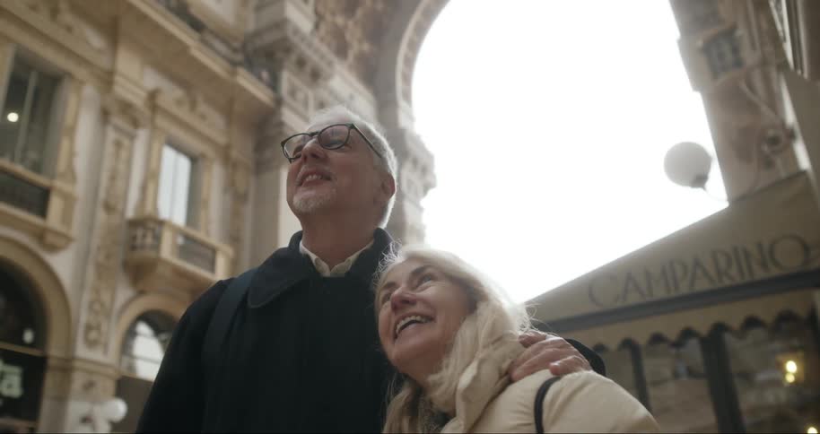 An elderly couple shares a joyful moment embracing as they admire the grand architecture of a historic shopping gallery bathed in soft natural light