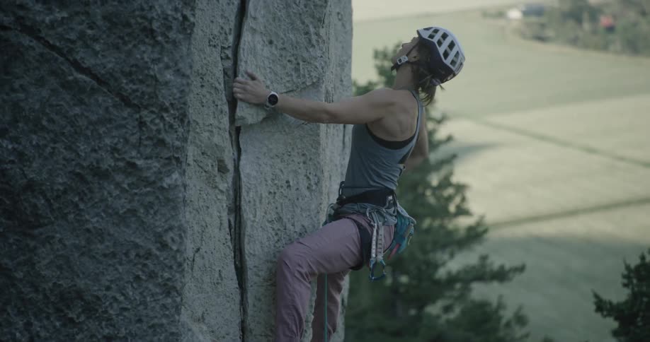 A female rock climber with climbing gear and a helmet carefully ascends a gray rock face