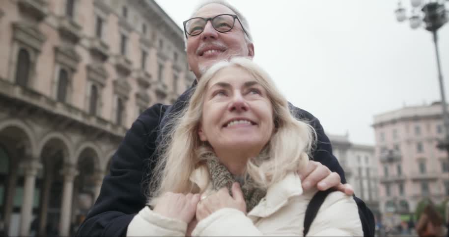 Elderly couple smile as they embrace in a square surrounded by historic arcaded buildings