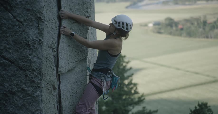 A female rock climber wearing a helmet and harness grips a crack in the rock face as she ascends with determination against a scenic countryside backdrop