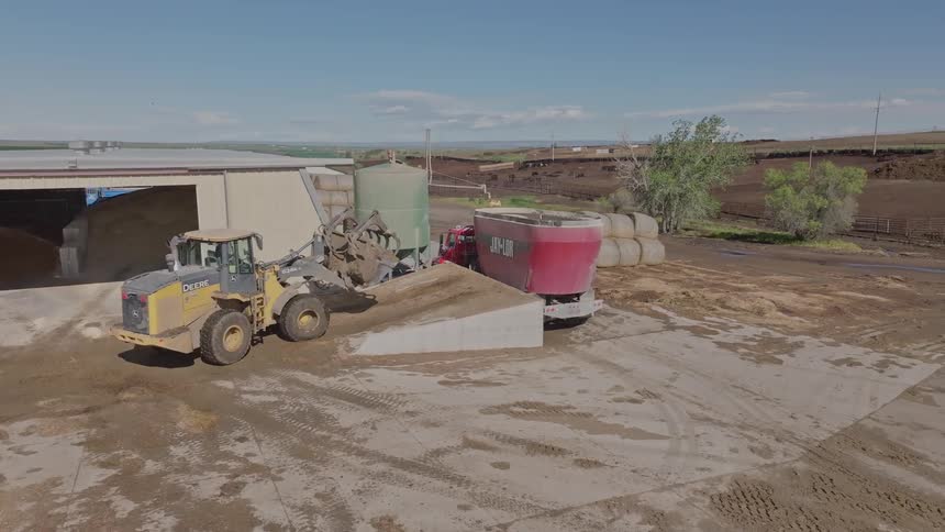 A yellow tractor loads feed into a red mixer truck at a farm with a barn hay bales and cattle visible in the background