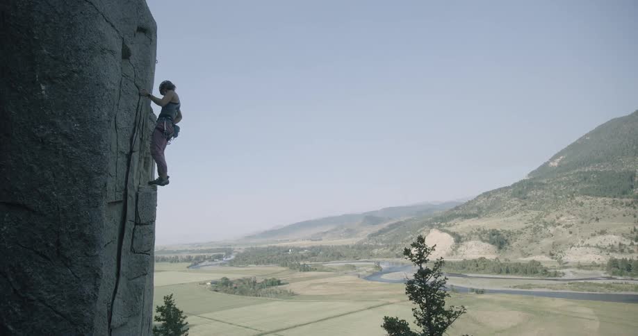 A rock climber ascends a sheer cliff face with a panoramic view of a valley and river sprawling below