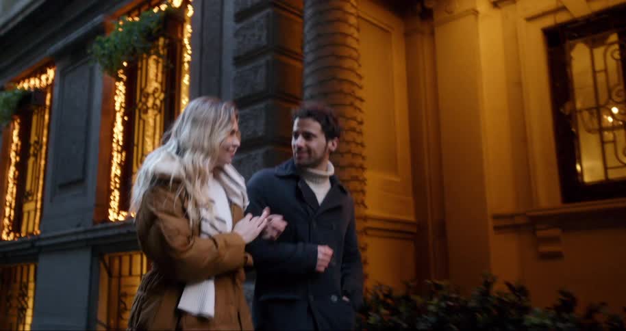 A young couple strolls arm in arm past a building adorned with warm glowing lights