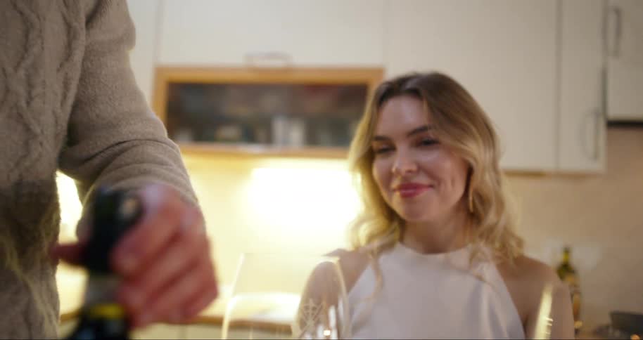 A man serves wine to a blonde woman seated at a kitchen table