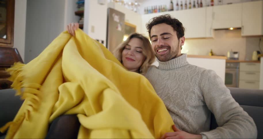 A couple is sitting on a couch in a cozy living room wrapped in a yellow blanket with a kitchen visible in the background