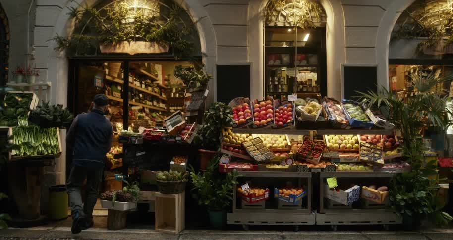 A man carrying some boxes into a store that has various fruits on an outside shelf