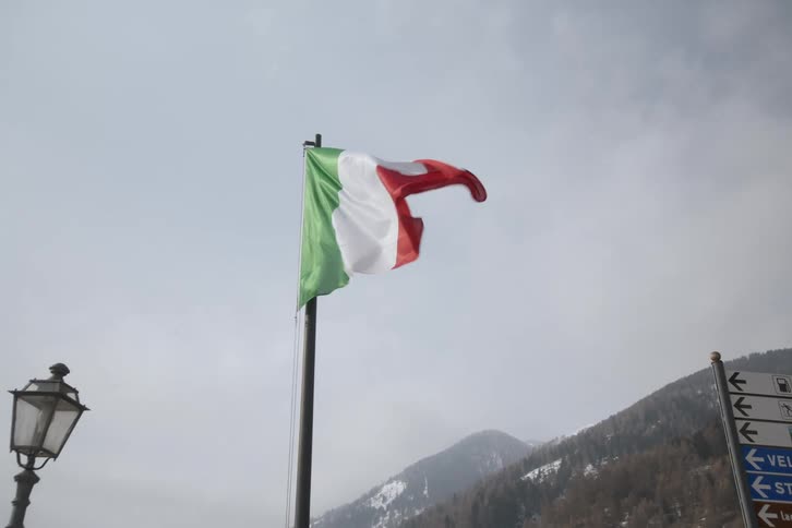 The italian flag waves against a backdrop of a cloudy sky and snow capped mountains